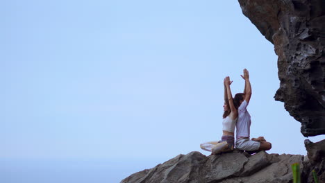 On-a-mountain's-peak,-a-man-and-woman-sit-back-to-back-on-a-rock,-engaging-in-meditation-and-yoga-with-the-ocean-as-their-backdrop